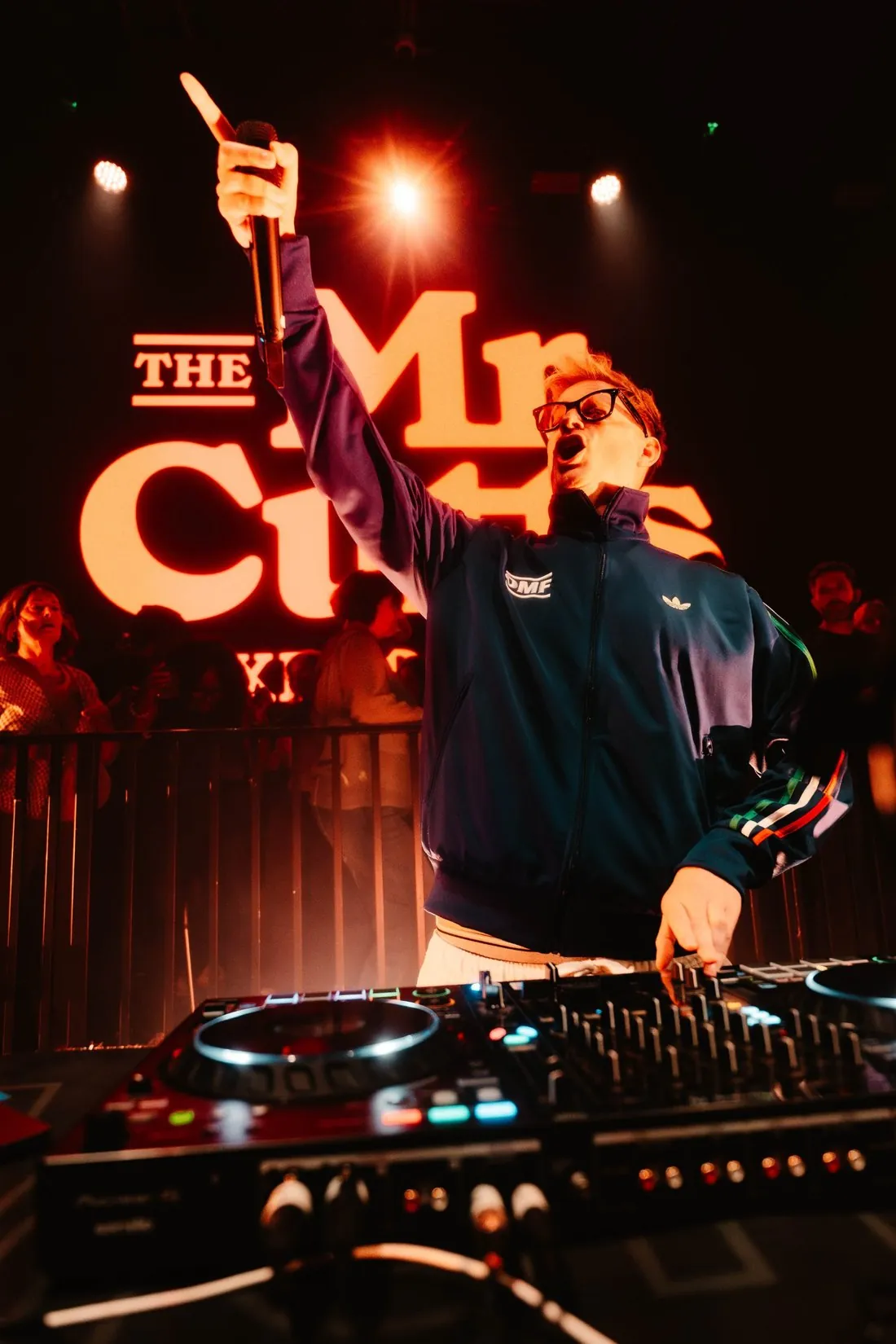 Owen Cutts with a microphone at the decks, The Mr Cutts Experience logo behind him